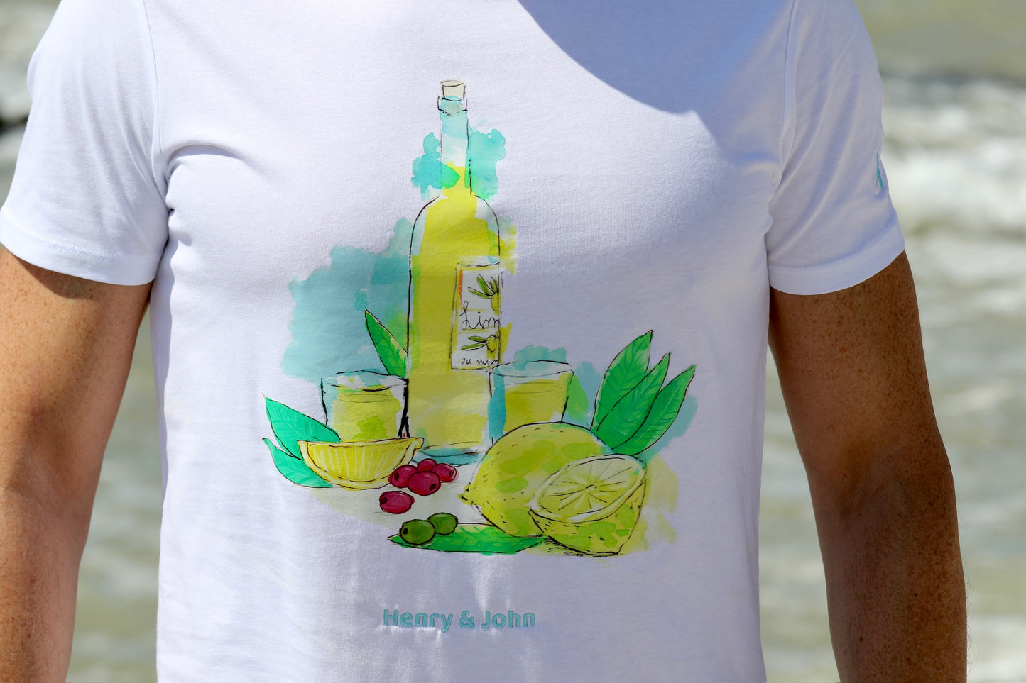 detalle de la camistea manga corta del limoncello de henry & john con la botella de limoncello, vasos llenos de la bebidaa y limones con sus hojas se ve el detalle bordado de la marca henry & john en hilo azul celeste