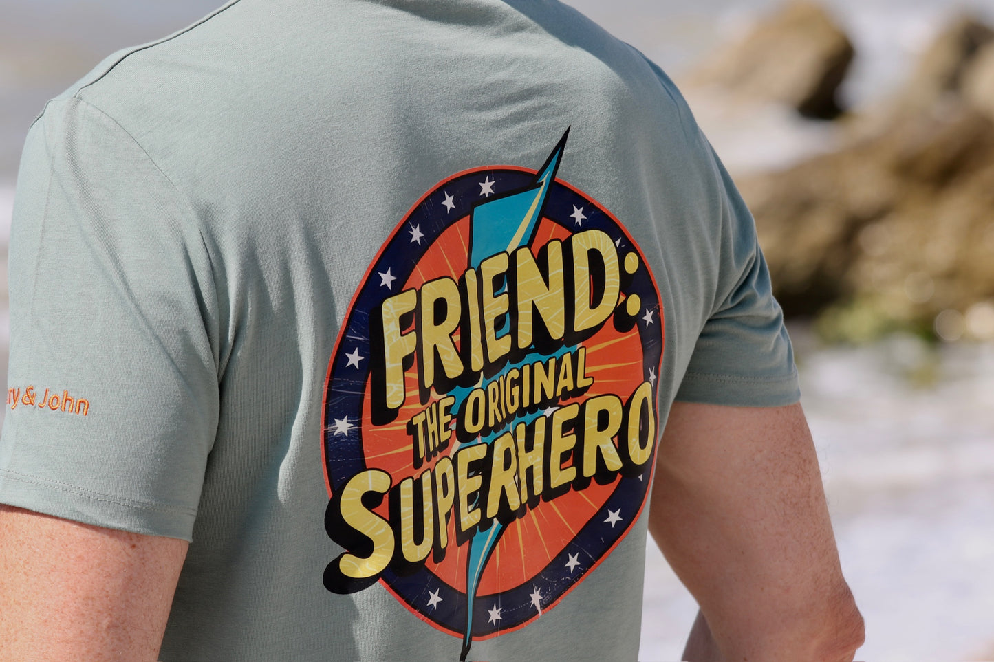 detalle lateral y dorsal de camiseta manga corta Friend: the original superhero con fondo difuminado en la orilla del mar