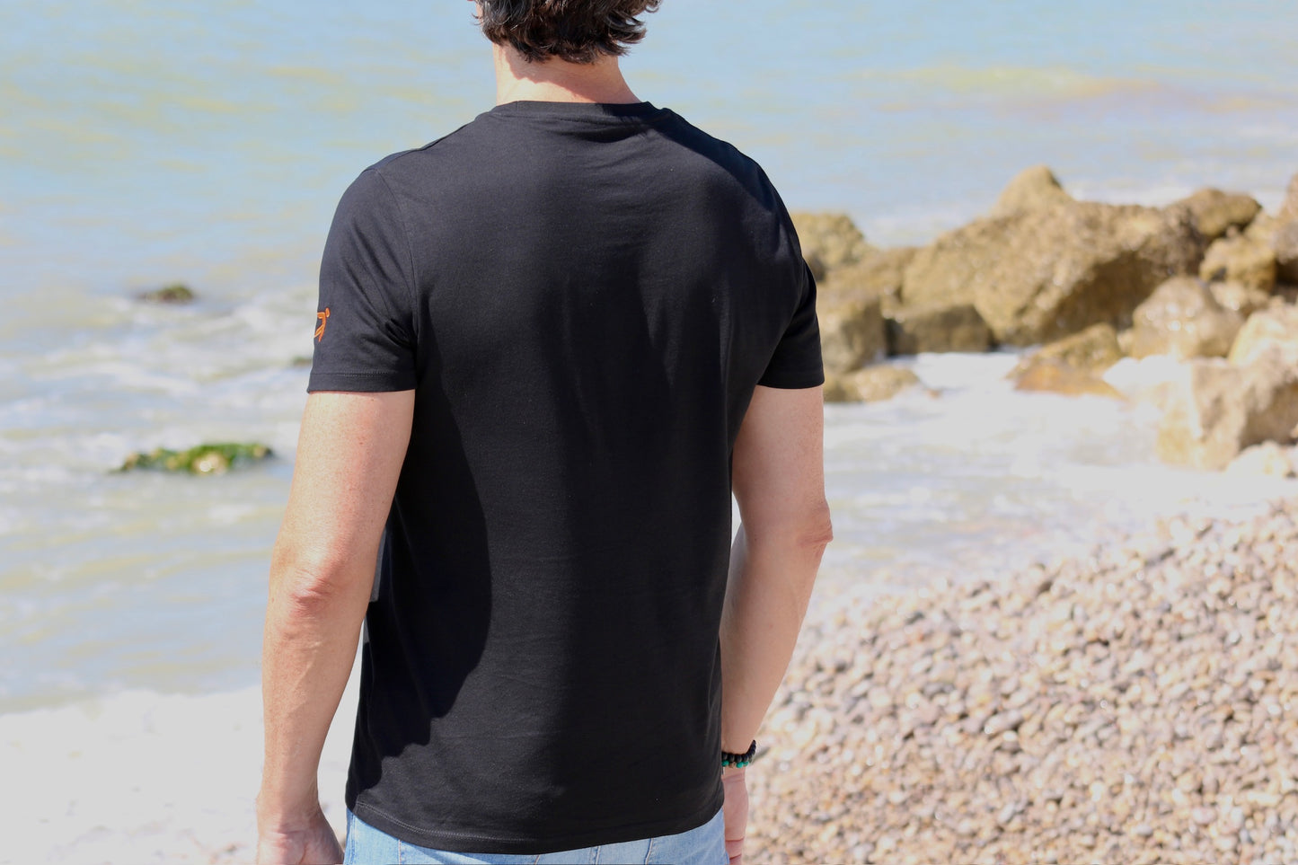 parte trasera con detalle en manga izquierda del logo de h&j de la camiseta manga corta negra algodo 100% orgánico de série enjoy your friends con fondo de mar y rocas difuminado