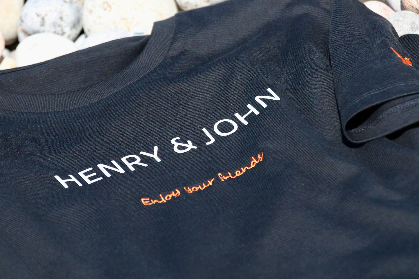 detalle de camiseta manga corta negra h&j de algodón 100% orgánico frontal bordado con letras blanca Henry & John y enjoy your friends en naranja y el detalles del logo de h&J bordado en color naranja en la manga izquierda doblada, la camiseta está sobre las piedras de la orilla del mar un poco difuminadas