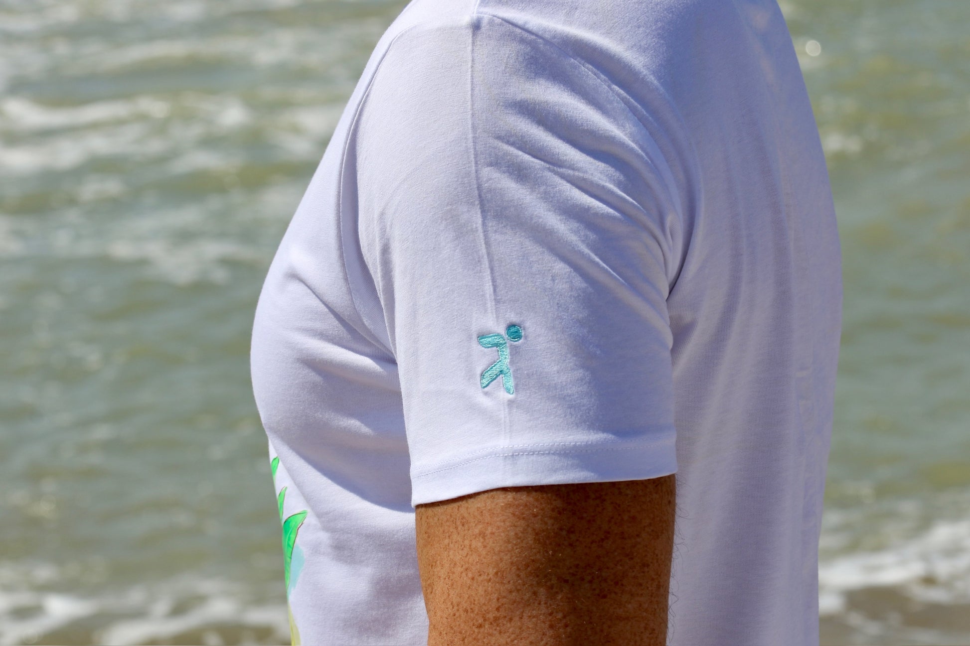 lateral de la camiseta blanca manga corta h&j limoncello con el logo en la manga izquierda bordado en hilo azul celeste de la marca henry and john figura abstracta de un atleta llegando a meta, se intuye el frontal de la camiesta en dtf en fondo difuminado de mar