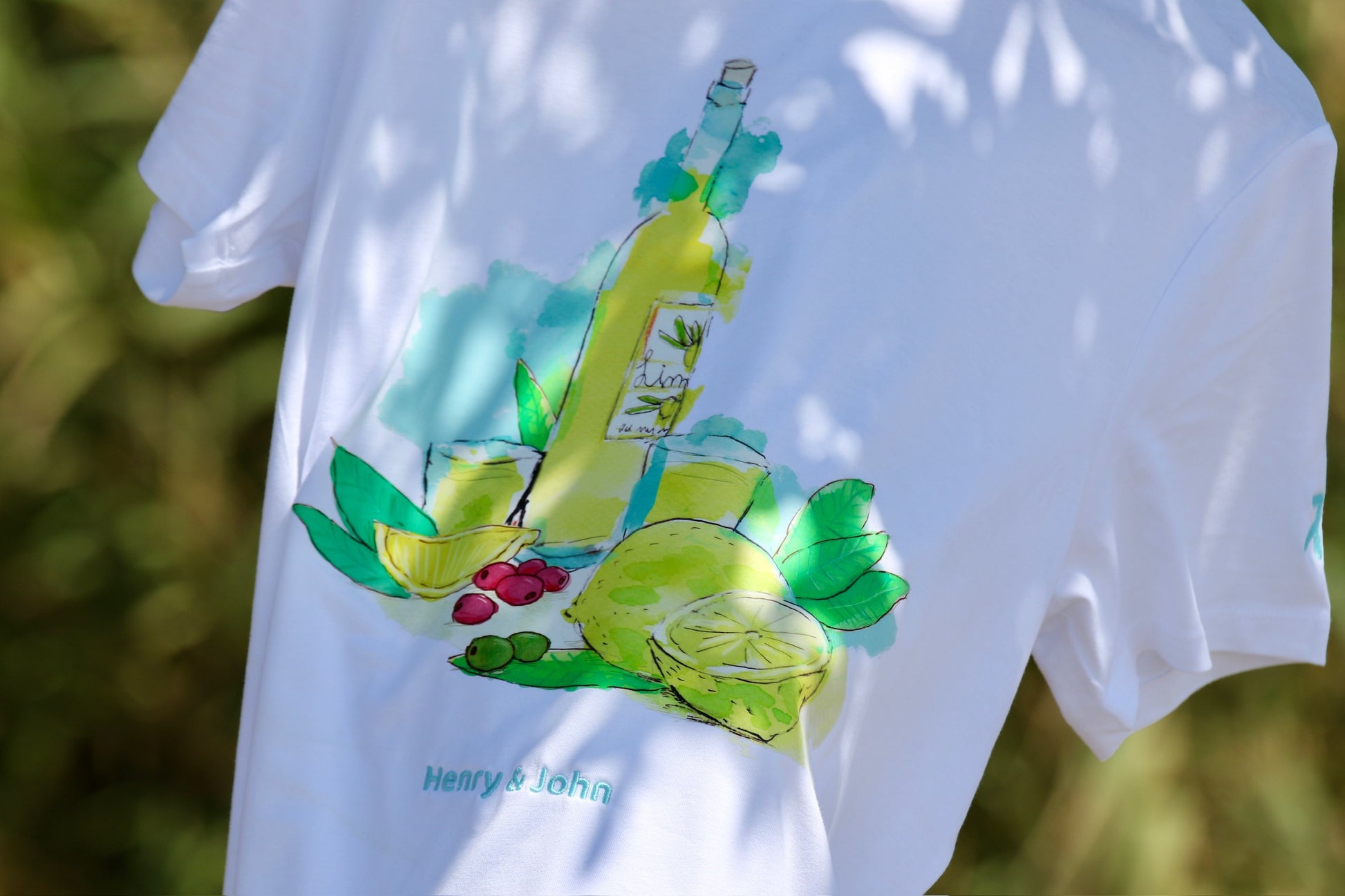 camiseta manga corta blanca limoncello de h&j sobre fondo de vegetación mediterranea se observa el logo bordado azul celeste henry and john en la parte baja del frontal de la camisesta, encima está el dtf del un motivo veraniegoque representa el frescor de beberte un limoncello con sus ingredientes los vasos y los limones, predomina los colores amarillos, verdes y azues  se intuye el logo de henry and john en la manga corta izquierda bordado en  hilo azul celeste.