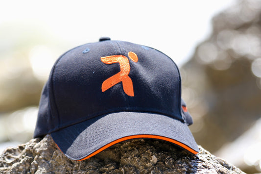 gorra de beisbol de color negra con logo de henry & John bordado con hilo de color naranja  en el centro, logo que representa un atleta abstracto llegando a meta se intuye el lateral izquierdo bordado en hilo naranja con el nombre de henry& John, la gorra esta encima de una roca y se ve el mar difuminado